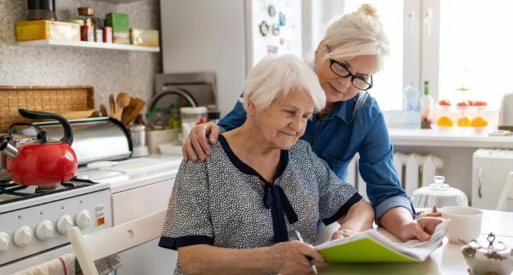 Older woman looks over elderly mother's shoulder in kitchen as they both look at folder on counter
