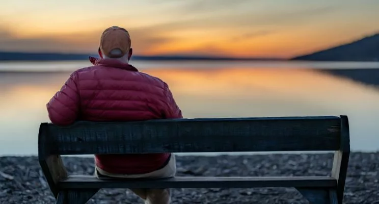 older man sitting on park bench overlooking a lake and watching the sunset